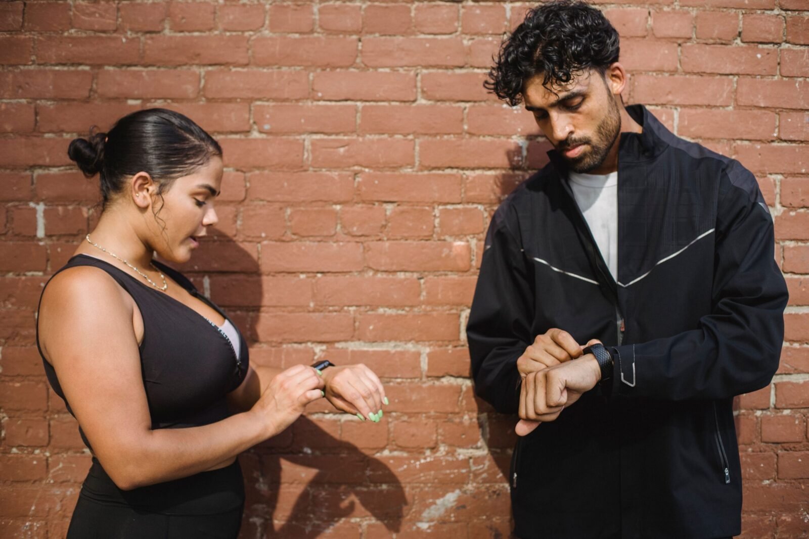 Two adults in sportswear checking their digital watches against a brick wall backdrop.